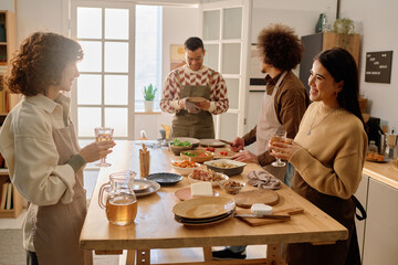 Old friends chatting lovely and holding crystal glasses with apple juice while biracial guys serving table to celebrate house-warming