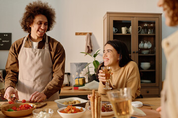 Brunette young adult woman leaning on table and holding glass full of apple juice while looking at her curly haired boyfriend with love-filled eyes