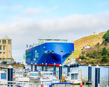 Pasajes, Spain - January 15 2025. A large hybrid-powered cargo ship docked in a harbor, accompanied by a tugboat, with a backdrop of lush hills and overcast skies.