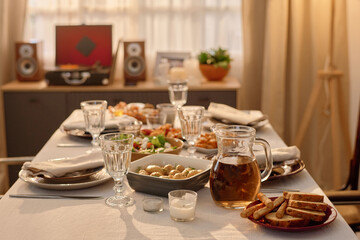 Medium close up of polished crystal glasses and ceramic plates with cotton napkins, jar with apple juice and homemade food on table covered with tablecloth