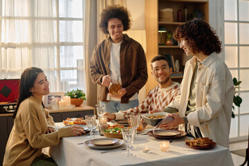Wide shot of biracial couple serving table with jar of apple juice and baking tray while their friends sitting at table