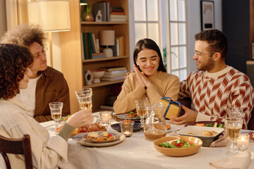 Young adult man patting his wife and giving small gift box wrapped in yellow paper and sating ribbon while their friends watching them