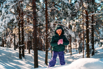 Naklejka premium Smiling child dressed in warm winter clothing is joyfully crafting a snowball in a snowy forest, embracing the fun of winter holidays amidst the stunning natural landscape