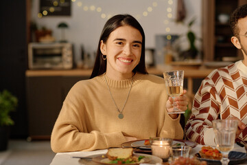 Smiling young adult brunette smiling widely and raising her glass with champagne while posing for portrait
