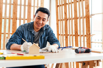 A man smiles while crafting a small wooden house model at a well-lit workspace, surrounded by tools and materials. Carpentry working at a home workshop studio.