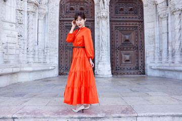 Naklejka premium A young beautiful asian woman with a beautiful long orange dress stands in front of beautiful architecture in Lisbon, Portugal. She pushes her hair back out of her face. She smiles at the camera.