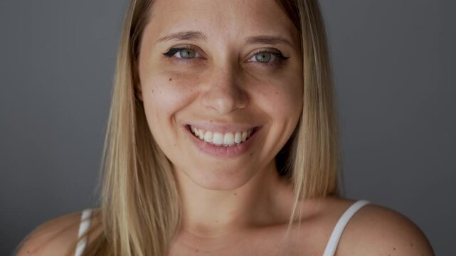 Young attractive caucasian blonde woman with green eyes and dimples on the cheeks turns her head and smiles sincerely on a dark grey background. Beautiful smile. Close-up
