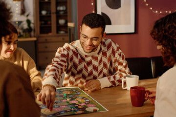 Fototapeta premium Company of friends sitting at wooden table looking at playing field and man making his game turn