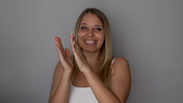 A young caucasian cheerful smiling blonde woman shows thumb up like gesture and claps her hands in assent and approval on a dark grey background. Applauding and congratulating girl