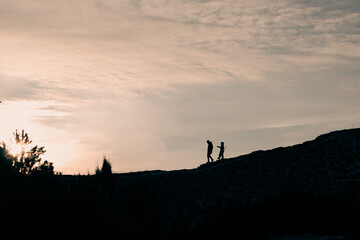 Fototapeta premium Contrasting silhouettes of two people in sunset light - an adult and a kid - on a hill in a park, forest. Family nature walk with a child. Adventure.