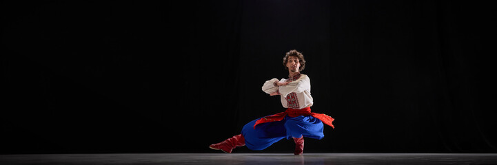 Single male dancer in low crouched pose, dressed in embroidered shirt, blue trousers, and red boots, showcasing focus and tradition against black background. Concept of traditions, art and history. Ad