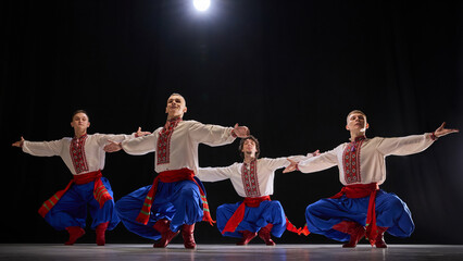 Group of male dancers performing synchronized jump, dressed in traditional embroidered shirts, blue trousers, and red boots, expressing traditions against black background. Concept of folk style