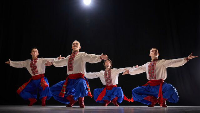 Group of male dancers performing synchronized jump, dressed in traditional embroidered shirts, blue trousers, and red boots, expressing traditions against black background. Concept of folk style