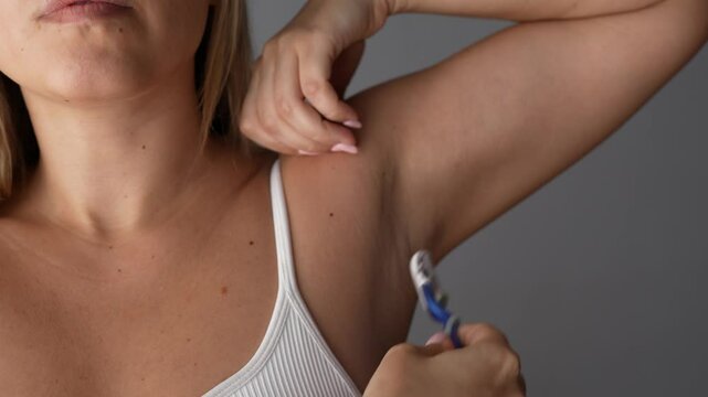 Young blonde woman shaving her armpit with a razor on a dark grey background. Getting rid of unwanted hair