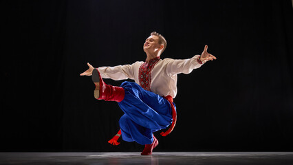 Male dancer holding balanced kick, arms wide for elegance, traditional attire emphasizing movement and heritage against black studio background. Concept of traditions, folk style, art and history. Ad