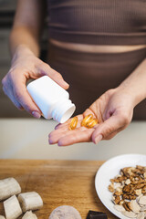 Athletic woman in sportswear is holding omega-3 capsules, fish oil supplements in her hand, in a kitchen