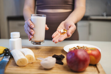 Athletic woman in sportswear holding a glass with a protein drink, milkshake, or smoothie and omega-3 capsules, fish oil supplement, white amino acid capsules or vitamins in her hand