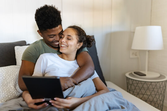 A cheerful couple cuddled together in bed, using a tablet to browse or watch content, enjoying a cozy and relaxing moment in a bright, modern bedroom