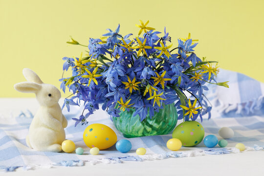 Easter still life, bouquet of spring blue and yellow flowers in a vase, bunny and eggs decor on the table. Beautiful postcard