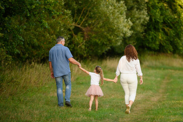 Obraz premium Family walking together in park on summer. Mom, dad, daughter spend time together walking along path holding hands, with backs to camera. Family, love and concept of parenthood, happy childhood.