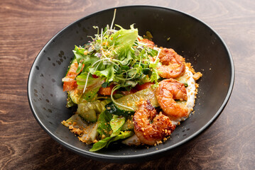 A vibrant dish featuring sautéed shrimp atop a bed of fresh greens and colorful vegetables, garnished with microgreens and sprinkled with crushed nuts. Served in a sleek black bowl.