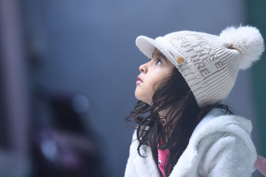 Side pose of an Asian Indian baby girl wearing winter woolen jacker and knitted cap looking up innocently.