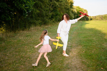 Fototapeta premium Woman and little girl play happily together with colorful toy airplanes on field. Mom and daughter run and have fun playing together on sunny day on weekend. Childhood concept, dreams, games.