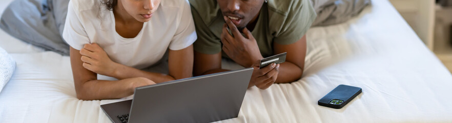 A young couple lying on their bed, shopping online with a tablet and credit card in hand, in a bright and cozy modern bedroom