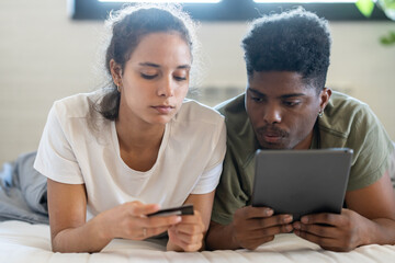 A young couple lying on their bed, shopping online with a tablet and credit card in hand, in a bright and cozy modern bedroom