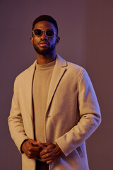 Fototapeta premium Stylish young man wearing a beige coat and sunglasses, showcasing confidence and modern fashion in a softly lit studio setting