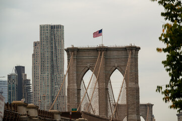 Fototapeta premium Brooklyn Bridge viewed from below on an overcast day with the American flag waving against a backdrop of city buildings