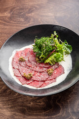 A gourmet dish featuring thinly sliced beef carpaccio arranged elegantly on a black plate, accompanied by a fresh green salad and a creamy dressing, set on a wooden background.