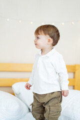 Fototapeta premium Child standing on bed in cozy room. A short-haired child in a white shirt and khaki pants explores a bright room filled with soft pillows.