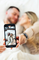 Couple sharing a moment in bed. A happy couple shares a cozy moment in bed while taking a selfie with a smartphone in their hands.