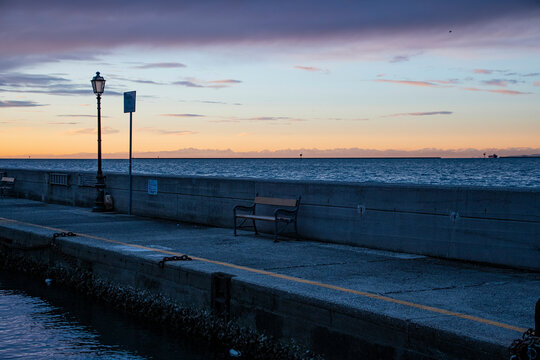 Muggia (Trieste, Italy): scenic view of  pier with a bench and a lamppost at dusk, blue sea water and orange and lilac sky