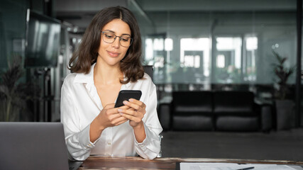 Focused Latin Hispanic female businesswoman holding smartphone sitting at desk in office. Successful middle age business woman manager ceo using cell phone mobile pc for online working. Copy space