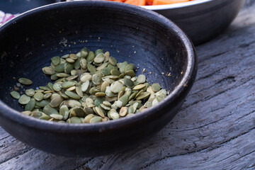 A close-up of a black rustic bowl filled with raw pumpkin seeds, placed on a weathered wooden surface. In the background, another bowl with sliced vegetables is partially visible.