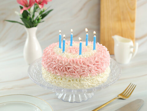 bolo de anivers&aacute;rio com velas azuis acesas e elementos ao redor do cen&aacute;rio como um vaso de flores, t&aacute;bua e jarra pequena branca 