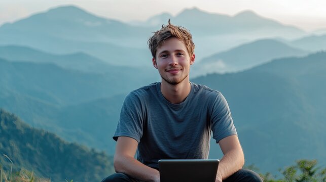 Young man smiling with laptop on mountain top, surrounded by ser