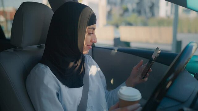 Young smiling Middle Eastern woman in hijab sitting in car and chatting online on smartphone. Female sipping hot drink from plastic cup. Enjoying warm beverage inside vehicle before going to work.