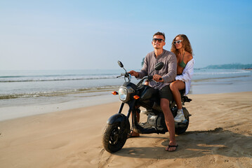 Obraz premium Couple rides motorbike on sandy beach near ocean. Man drives motorcycle, woman sits behind smiling. Enjoying adventure travel, seaside freedom, summer vibes.
