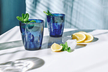 Summer refreshing lemonade drink with mint, ice and lemon slices on white surface and blue background. Fresh healthy cold lemon beverage. Infused water with lemon.