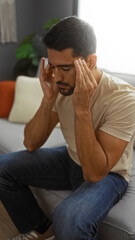 Obraz premium Young man with beard sitting on couch in living room with hands on temples looking stressed indoors at home with natural light from window