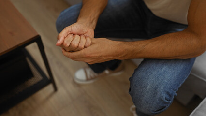Young hispanic man seated indoors with hands clasped, wearing casual jeans in a modern apartment living room setting, implying a relaxed home atmosphere.