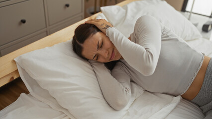 Young woman covering her ears with hands while lying on a bed in a bedroom of a house indoors