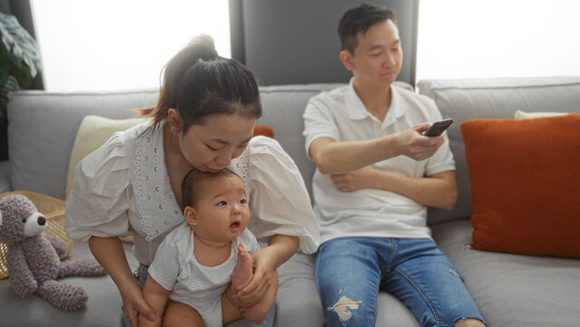 Mother and father with baby boy sit in living room; mother kisses baby's head while father holds remote on couch, creating a warm family moment indoors.