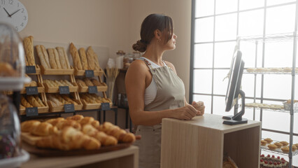 Woman standing in bakery wearing apron beside counter with breads and pastries displayed in the...