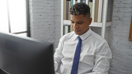 Young hispanic man working on a computer in an office setting, dressed in formal attire, focused and professional with a modern interior background