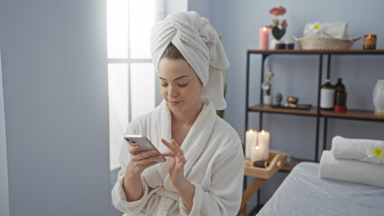 Young woman at spa wearing towel on head and robe, using phone in wellness center's calming interior
