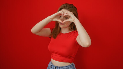 Young woman forming a heart shape with her hands in front of a red wall, wearing a red top and blue jeans, isolated against the vibrant background, creating an attractive and cheerful scene